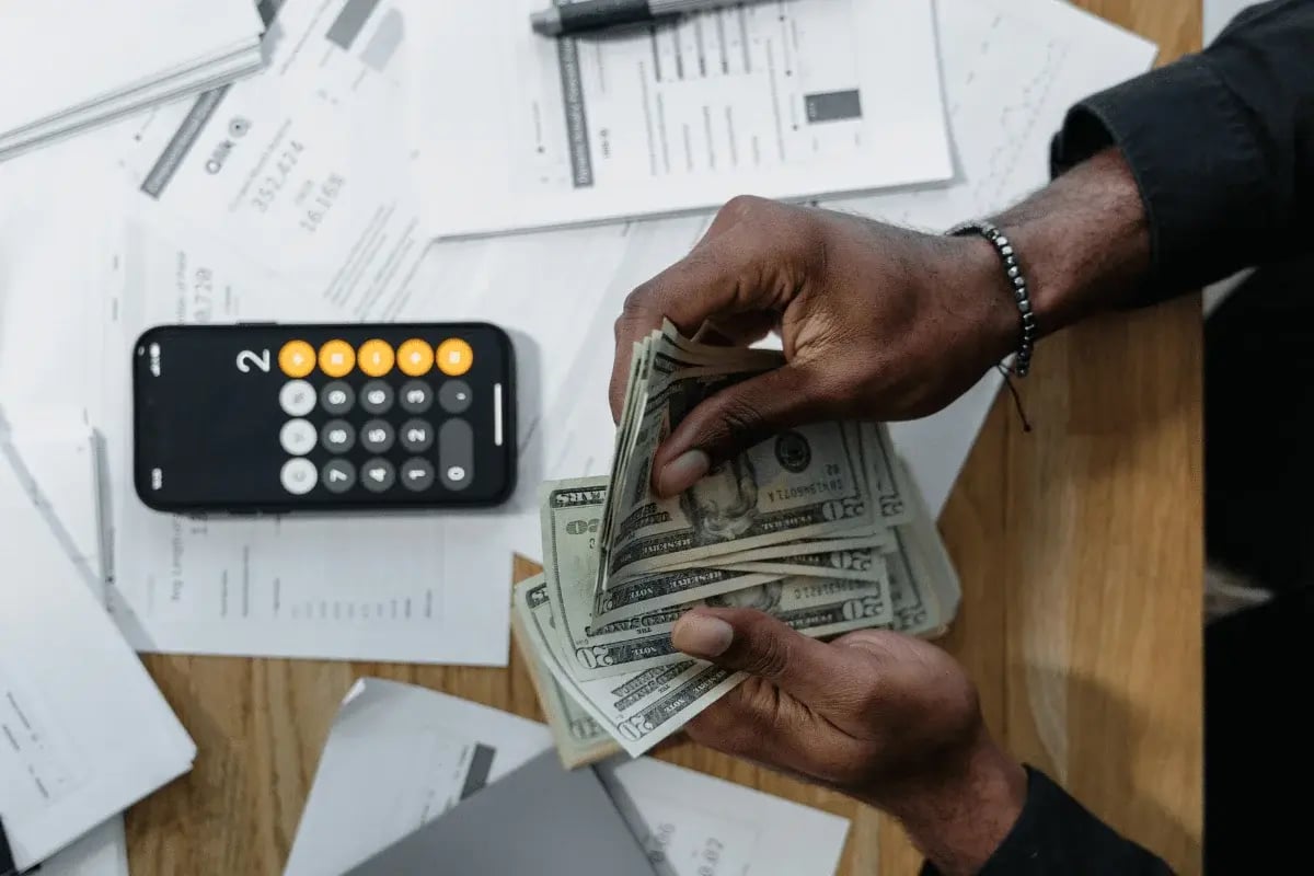 Close-up of man counting cash with phone calculator on desk.