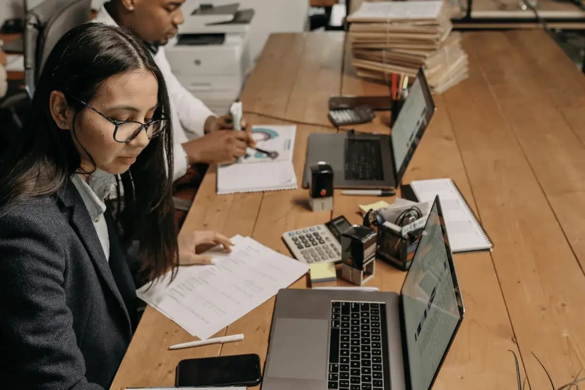 A close-up of a woman analyzing paperwork while another person works on a laptop, with a variety of office tools like calculators and stamps.