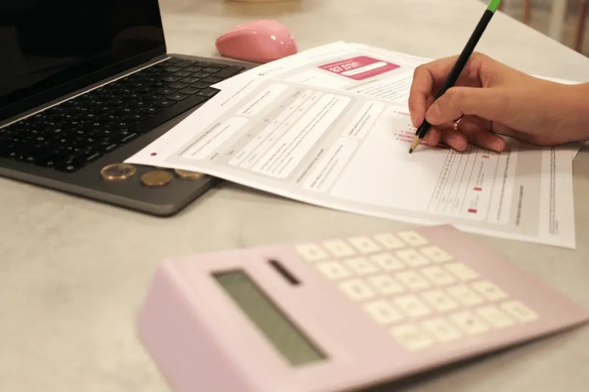 Hand writing on finance papers beside laptop and coins on desk.