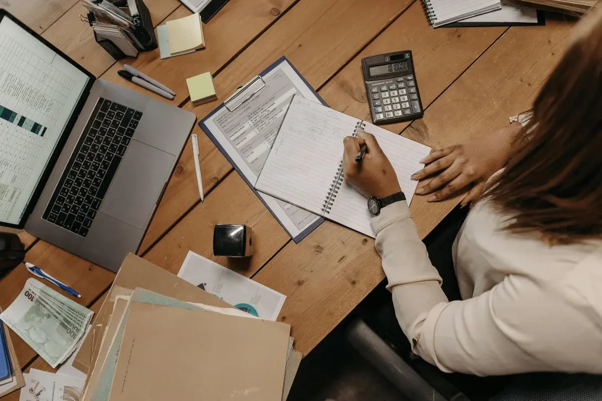 A person writes in a notebook at a wooden desk with a laptop, documents, calculator, and folders, conveying a busy and organized work environment.