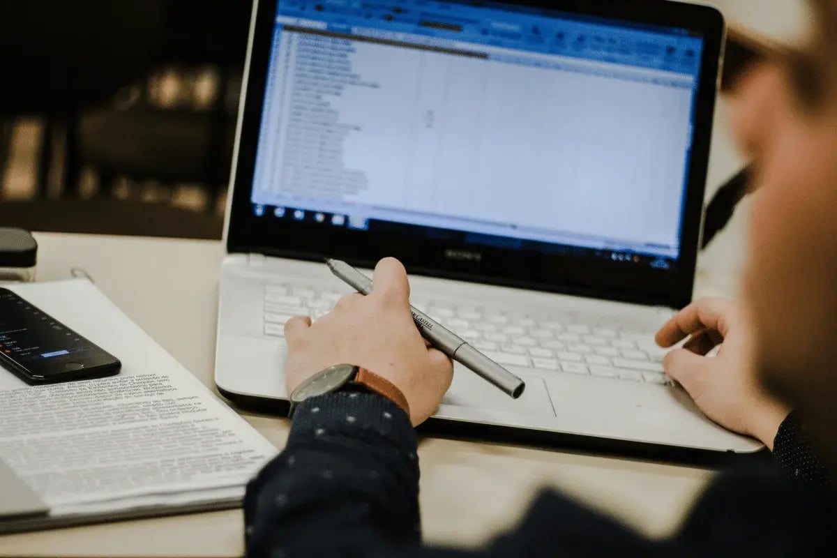 Hand holding pen while reviewing business reports on computer.