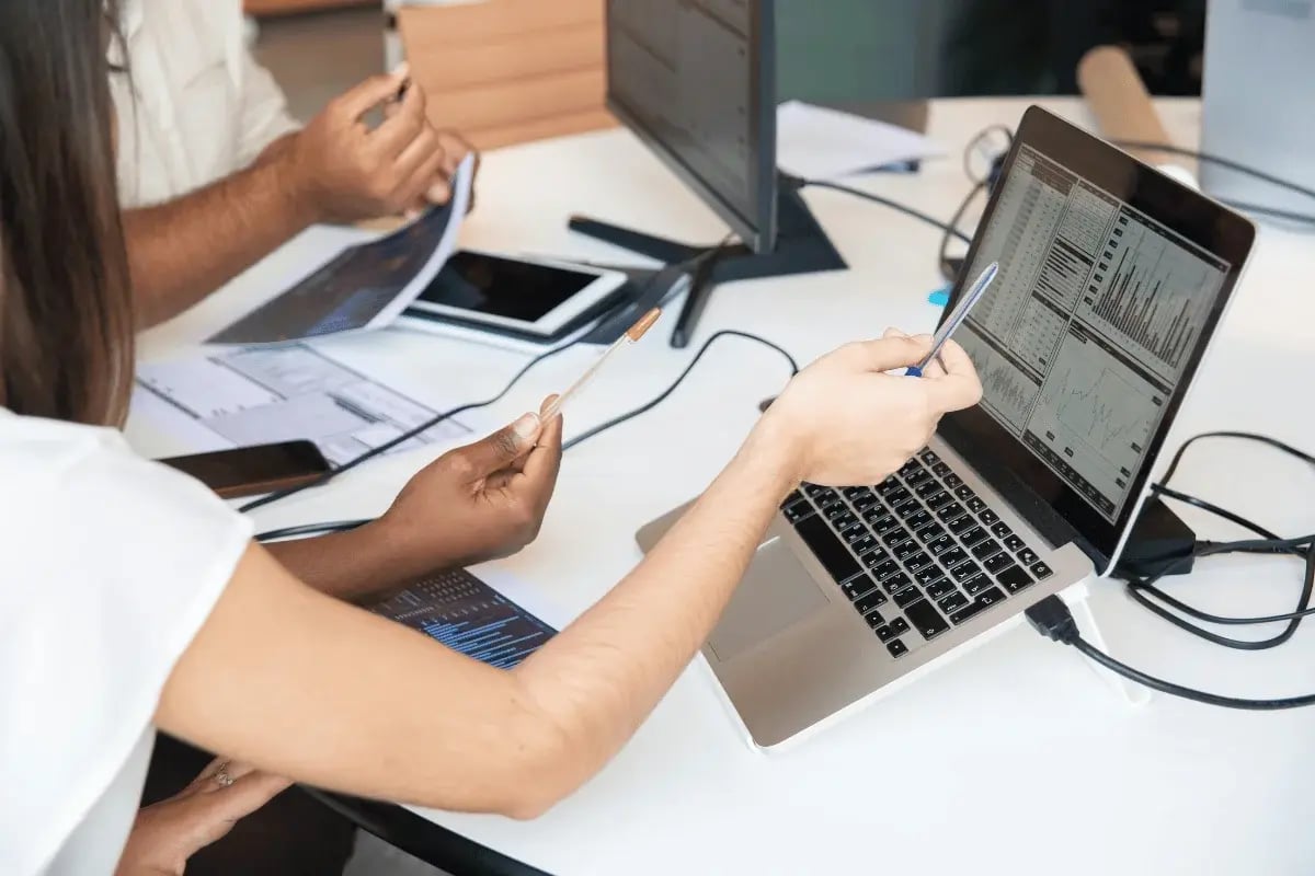 Two people in a meeting, analyzing data on a laptop, while reviewing printed reports and using smartphones.