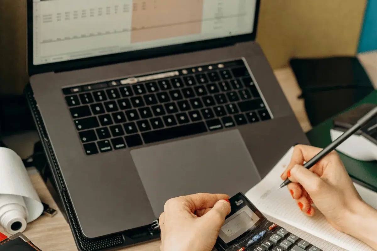 Person using a calculator while taking notes beside a laptop.