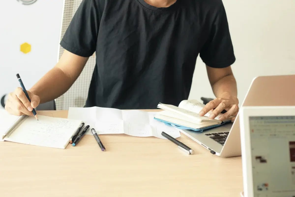 Student writing notes while referencing a book and laptop.