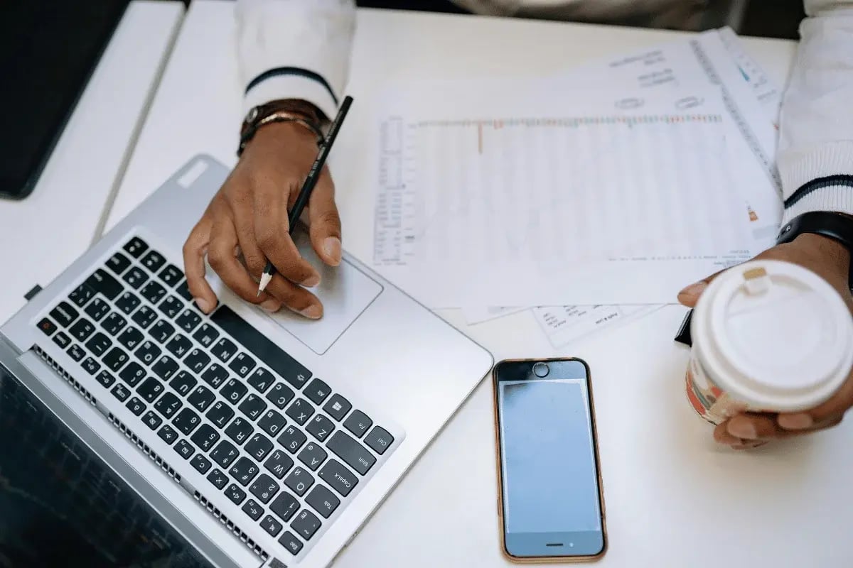 An individual at a white desk works on a laptop, holding a pen over charts. A smartphone and a coffee cup are nearby, suggesting a busy workspace.