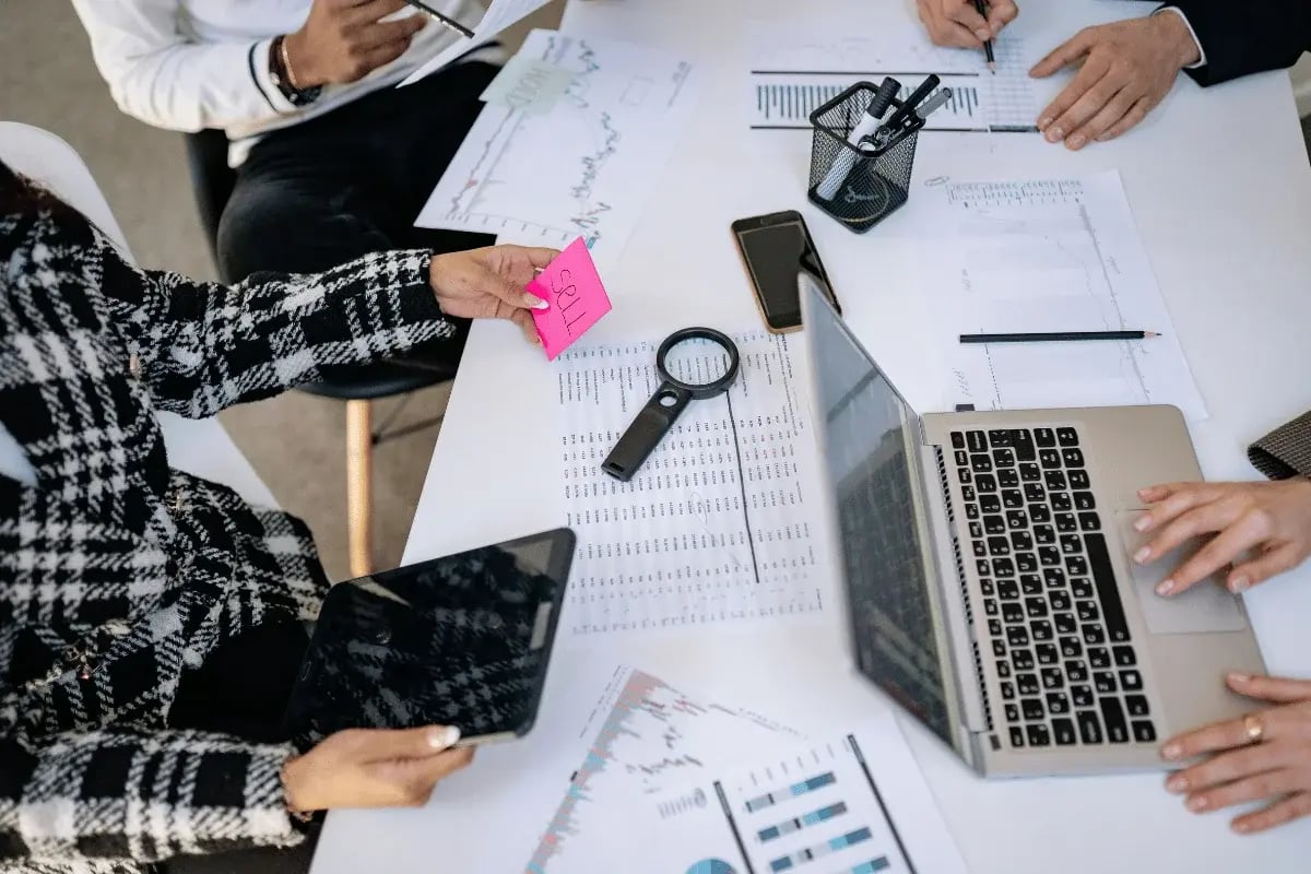 People collaborate at a table with documents, graphs, and a laptop. A magnifying glass and smartphone are present, conveying teamwork and focus.
