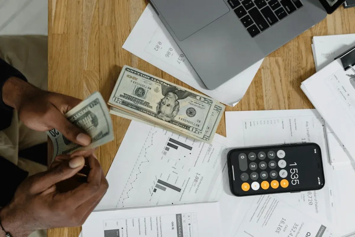 Close-up of someone counting cash with dollar bills on a desk, surrounded by papers, a calculator, and a laptop.