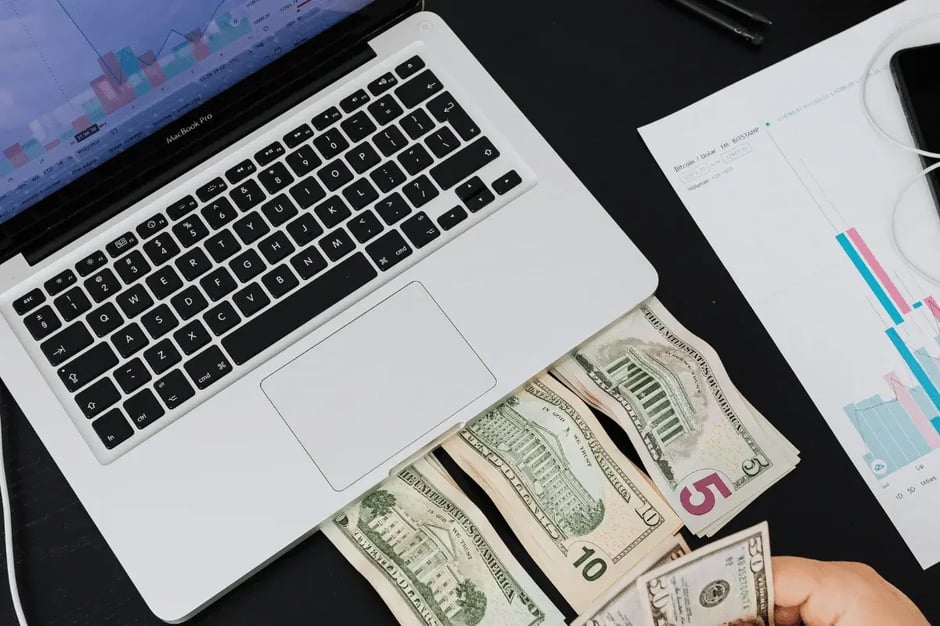 Cash and currency notes placed beneath a MacBook with trading charts on screen.