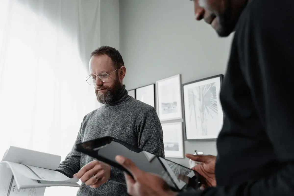 A bearded man in a gray sweater is holding financial documents while another person uses a tablet in a contemporary office space.