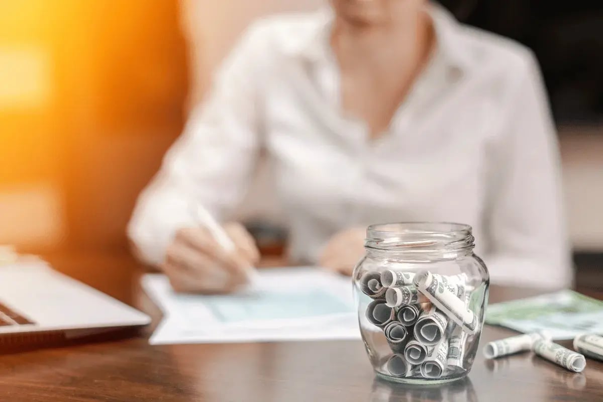 Woman writing at desk with money jar in foreground.