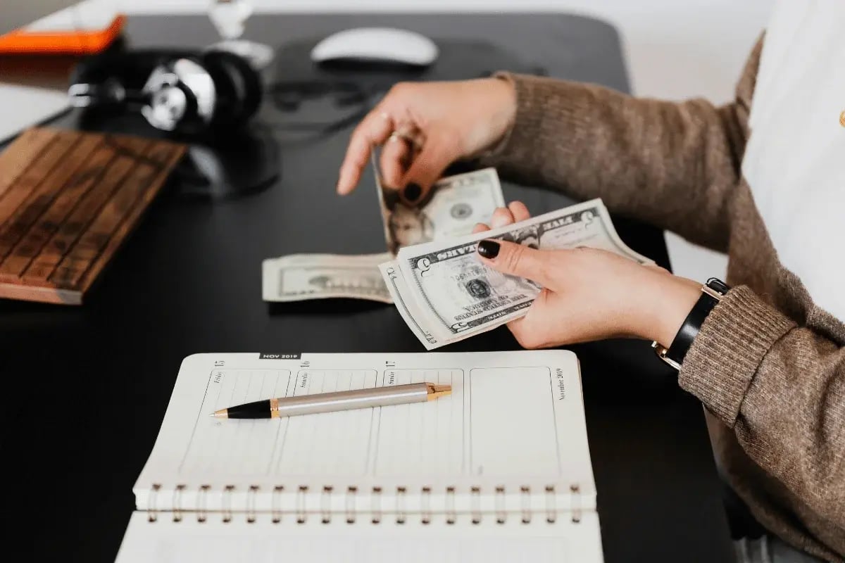 A person handling cash on a desk with a calendar notebook, pen, and a wooden keyboard in the background.