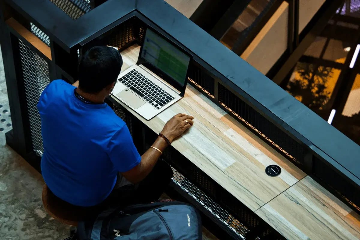 Remote worker typing on a laptop at a wooden desk in a public area.