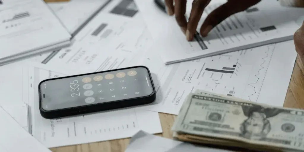 A smartphone displaying a calculator app rests on a cluttered desk with financial documents and cash, indicating budgeting or calculations.